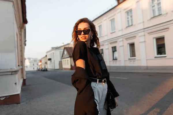 Joven mujer europea bastante hipster en un elegante abrigo largo negro en vaqueros blancos en una camiseta posando de pie en la ciudad cerca de los edificios blancos vintage. Elegante modelo de chica disfruta del sol . — Foto de Stock