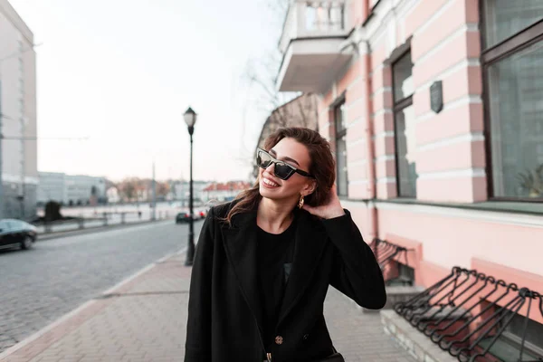 Mujer hipster bastante joven positiva en abrigo negro vintage con gafas de sol elegantes en camiseta de moda camina por la calle y disfruta de un cálido día de primavera. Linda chica alegre se relaja al aire libre. Estilo juvenil . — Foto de Stock