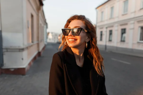 Jonge vrolijke hippe vrouw met een positieve glimlach in zwarte zonnebril in een stijlvolle jas poseren in de buurt van een wit vintage gebouw in de stad op een heldere zonnige dag. Joyful meisje model geniet van de zonsondergang. — Stockfoto