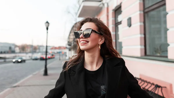 Досить кумедний хіпстер молода жінка в темних стильних сонцезахисних окулярах в модному чорному пальто в футболці йде по вулиці біля старовинної будівлі і позитивно посміхається. — стокове фото