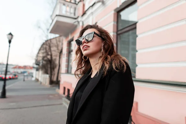 Jolie belle jeune femme à la mode en lunettes de soleil élégantes dans un manteau noir vintage posant dans la rue près d'un bâtiment rose. Urban European positive girl voyage autour de la ville . — Photo