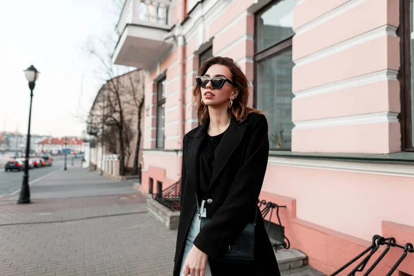 Modieuze moderne jonge vrouw hipster in een zwarte elegante jas in een T-shirt in jeans in een stijlvolle zonnebril wandelen langs de straat in de buurt van vintage gebouwen. Europees meisje reist door de stad. Voorjaar — Stockfoto