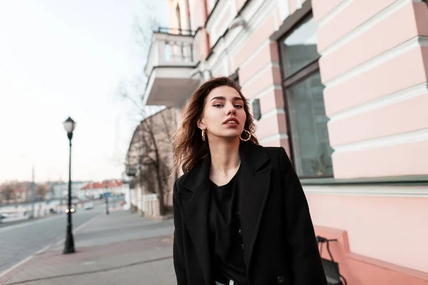 Elegante mujer joven atractiva en un abrigo negro con estilo en una camiseta con pendientes de oro está de pie al aire libre cerca de edificios antiguos. Chica moderna en un paseo en el día de primavera. Moda ropa de mujer . — Foto de Stock