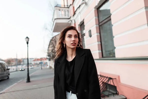 ジーンズのファッショナブルなTシャツでシックな黒いコートで自然なメイクアップを持つ現代の魅力的な若い女性は、市内のヴィンテージビルの近くにポーズをとります。屋外で美しいヨーロッパの女の子のモデル。ファッション. — ストック写真