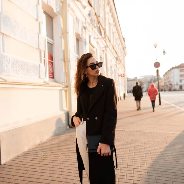 Mujer moderna y hermosa hipster en un abrigo elegante en una camiseta en jeans blancos con un bolso de cuero en gafas de sol posando cerca de un edificio vintage al aire libre. Chica atractiva disfruta de un día soleado. Juventud . — Foto de Stock