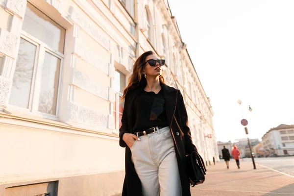 Mujer hipster joven bastante moderna en jeans de moda con un bolso de cuero en gafas de sol oscuras en un abrigo largo negro caminando por las calles de la ciudad cerca de un edificio vintage en un día soleado. Chica con estilo . — Foto de Stock