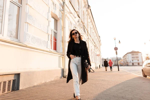 Belle jeune femme hipster en lunettes de soleil sombres dans un manteau noir élégant dans un T-shirt en jeans avec un sac à main se déplace le long de la rue près d'un bâtiment vintage par une journée ensoleillée. Jolie fille . — Photo