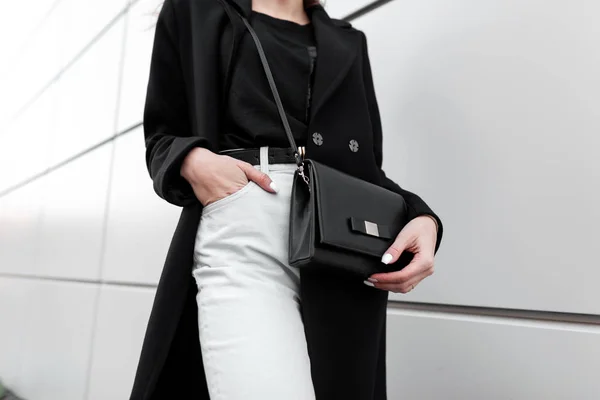 レザースタイリッシュなブラックのハンドバッグを持つ黒のトレンディーなTシャツのヴィンテージホワイトジーンズのファッショナブルな長い黒のコートの若いスタイリッシュな女性が屋外に立っています。現代の女性のファッション。春-秋. — ストック写真