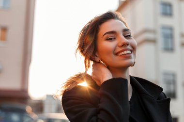 Modaya uygun siyah paltolu siyah vintage tişörtlü oldukça komik genç kadın, binaların yakınındaki şehirde turuncu parlak bir gün batımının keyfini çıkarıyor. Pozitif sevimli kız modeli gülümsüyor ve güneşte rahatlatır.