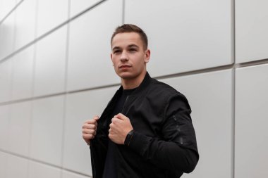 Şık bir saç modeli ile siyah moda ceket çekici yakışıklı genç adam şehirde sokakta gri modern duvar yakınında duruyor. Avrupalı bir adam. Modaerkek giyim.