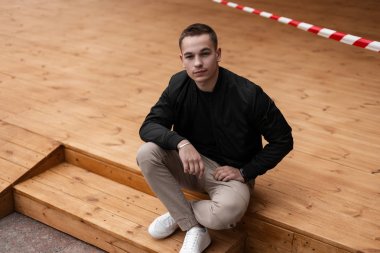 Modaya uygun siyah ceketli vintage pantolonlu şık spor ayakkabılı şehirli genç adam, ilkbahar da şehirdeki bir yaz kafenin ahşap terasında oturuyor. Çekici modern adam hafta sonu sahiptir.