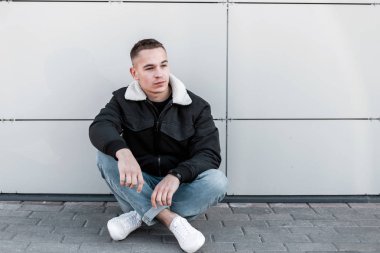 Şehirde modern bir binanın yakınında oturan rahatlatıcı bir saç modeli ile beyaz spor ayakkabı şık bir ceket şık kot Pretty genç adam. Şık adam sıcak bahar gününde açık havada dinleniyor.