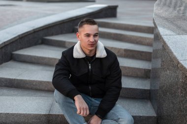 Mavi vintage kot şık bir siyah ceket şık bir saç modeli ile Çekici neşeli genç adam ilkbahar gününde açık havada oturur. Yakışıklı modern adam modeli geri kalanı sahiptir.