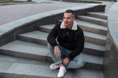 Şık ceketli, spor ayakkabılı, kot pantolonlu, neşeli genç adam şehirde klasik taş bir merdivende oturuyor ve gülümsüyor. Neşeli adam modeli dışarıda dinleniyor. Modaya uygun erkek kıyafetleri..