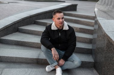 Siyah moda ceket deri moda spor ayakkabı şık kot Pretty sevimli genç adam açık havada bir vintage taş merdiven üzerinde oturan dinleniyor. Kentsel adam bir bahar gününde şehirde rahatlatır.