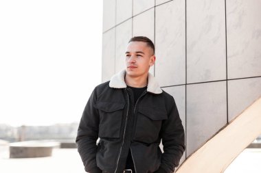 Şık bir saç modeli ile şık bir t-shirt bir vintage siyah ceket güzel çekici genç adam parlak bir bahar gününde modern binanın yakınında pozlar. Şehirli erkek modeli. Şık erkek giyim.