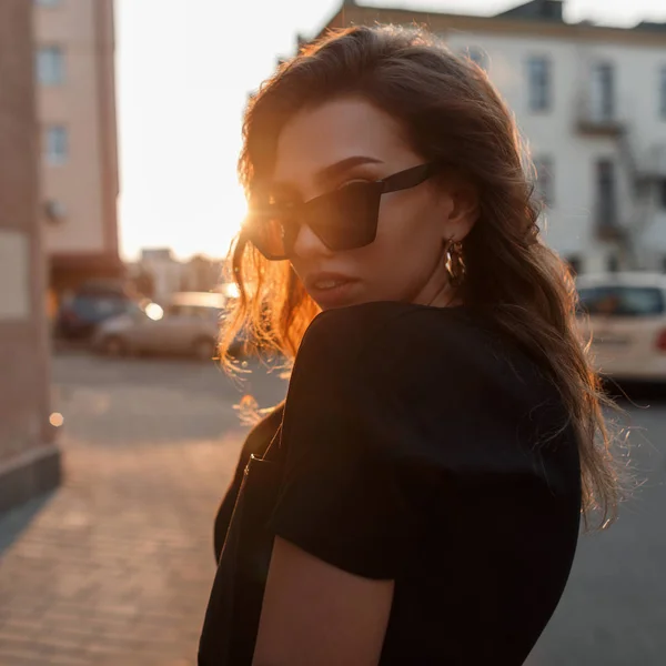 Портрет сучасної красивої молодої хіпстерки в стильних сонцезахисних окулярах в чорній модній футболці на тлі яскравого сонячного світла. Приваблива міська дівчина на прогулянці в сонячний день . — стокове фото