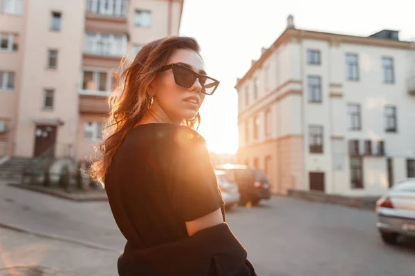 Європейський хіпстер молода жінка в старовинних темних сонцезахисних окулярів в чорній футболці з пальто прогулянки по місту на фоні яскравого помаранчевого заходу сонця. Стильна дівчина любить сонце. Задоволення. — стокове фото