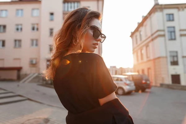 Jolie jeune femme hipster dans des lunettes de soleil à la mode dans un t-shirt noir dans un manteau voyage à travers la ville sur le fond d'un coucher de soleil orange vif. Urban girl bénéficie d'une promenade dans la soirée de printemps . — Photo