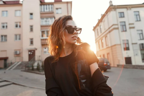 Молодая городская современная хипстерша в черной стильной футболке в темных модных солнцезащитных очках позирует на улице возле домов в городе. Красивая модель девушки наслаждается ярким оранжевым солнцем. Добрый вечер. . — стоковое фото