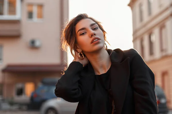 Jeune jolie femme dans un t-shirt élégant noir dans un manteau vintage avec maquillage naturel avec des lèvres sexy près de maisons vintage dans la ville. Fille attrayante bénéficie d'un soleil orange vif . — Photo