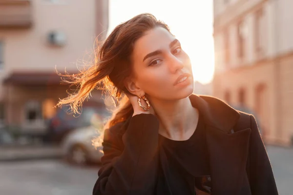 Portrait d'une jeune femme mignonne attrayante avec une coiffure élégante avec de beaux yeux avec des lèvres sexy en vêtements noirs à la mode sur un fond de coucher de soleil de printemps orange vif. Élégante fille se détend . — Photo