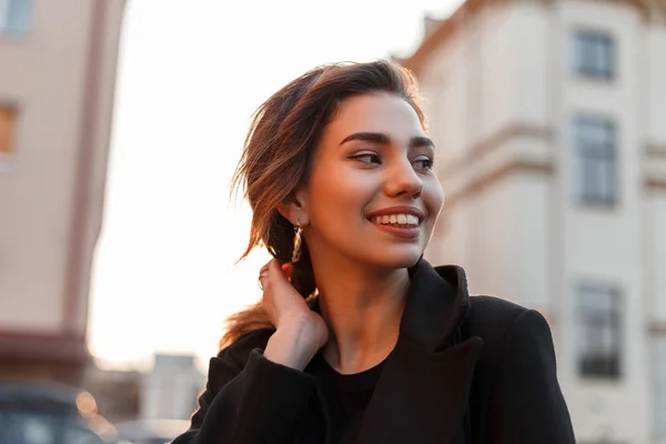 Portrait d'une jolie jeune femme drôle et heureuse avec un sourire mignon dans un manteau noir à la mode à l'extérieur de la ville par une journée ensoleillée. Joyeuse modèle de fille élégant bénéficie week-end ensoleillé . — Photo