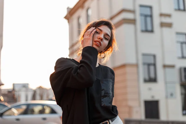 Mujer joven con estilo bastante moderno con maquillaje natural con labios sexy en una camiseta de moda en un abrigo de moda cerca de los edificios antiguos al aire libre. Linda chica disfruta de la luz del sol naranja brillante . — Foto de Stock