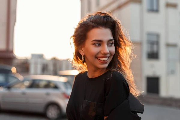 Lycklig ganska positiv ung kvinna i en snygg T-shirt i svart päls står och leende nära Vintage byggnader på en bakgrund av ljusa orange våren solnedgång. Joyful Girl Model njuter av solskenet. — Stockfoto