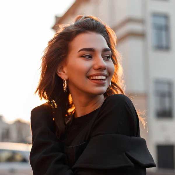 Joyful engraçado jovem mulher em uma camiseta vintage preto com brincos dourados goza de um pôr do sol laranja brilhante em pé na cidade perto de edifícios. Menina atraente positiva sorri e relaxa ao sol . — Fotografia de Stock