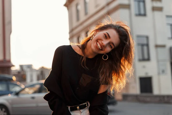 Lustige positive junge Frau in stylischer schwarzer Kleidung posiert und lächelt positiv in der Stadt vor einem frühlingshaft leuchtend orangefarbenen Sonnenuntergang. attraktives fröhliches Mädchen reist und genießt die Sonne. — Stockfoto