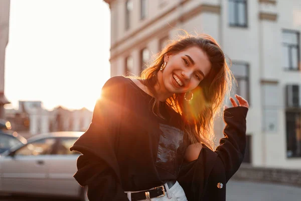 Красива досить позитивна молода жінка в стильній футболці в білих джинсах стоїть і сміється біля старовинних будівель на тлі яскраво-оранжевого весняного заходу сонця. Щаслива дівчина насолоджується сонячним світлом . — стокове фото
