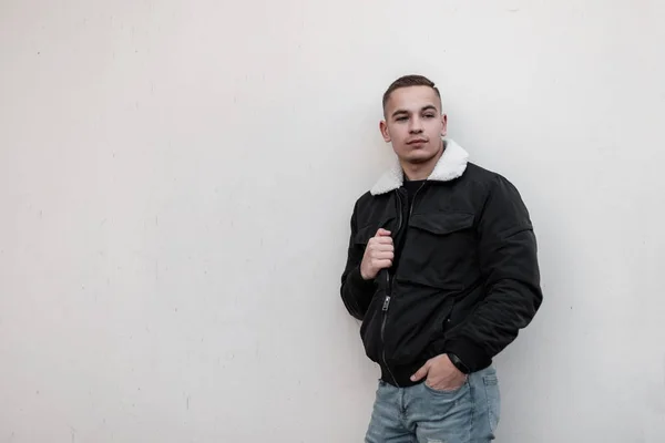 Attractive cute young man in blue stylish jeans in a fashionable jacket with white fur with a hairstyle rests standing near a wall outdoors in the city. European guy. Trendy menswear.