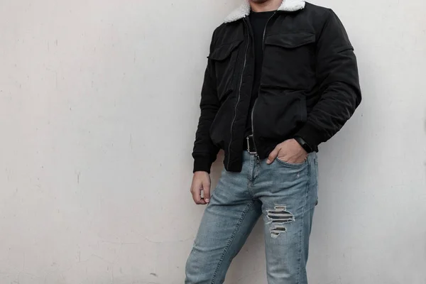 Jovem moderno na moda em uma jaqueta elegante preta com um colarinho branco em jeans de moda azul em uma camiseta preta elegante posando perto de um edifício moderno. Nova coleção de roupas masculinas.Close-up — Fotografia de Stock