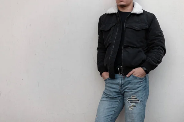 Jonge stijlvolle man in een zwart trendy jasje met een witte kraag in vintage blauwe jeans in een zwart t-shirt staat vlak bij de grijze muur. Modieuze heren bovenkleding. Straatstijl. Close-up van een mannelijk lichaam. — Stockfoto