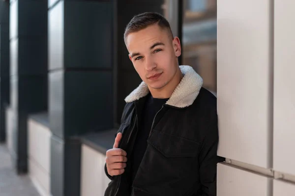 Jovem legal com um penteado na moda em uma jaqueta preta na moda em uma camiseta posando de pé perto da janela de um edifício moderno na cidade. Um tipo porreiro na rua. Roupas masculinas elegantes . — Fotografia de Stock