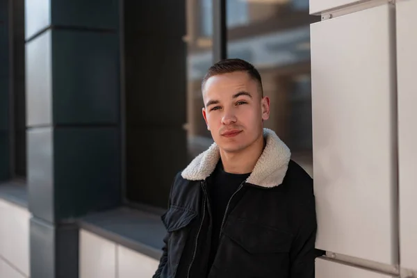 Bonito jovem com um penteado elegante em uma jaqueta preta na moda em uma camiseta posando de pé perto da janela de um edifício moderno na cidade. Um tipo urbano ao ar livre. Roupas masculinas na moda . — Fotografia de Stock