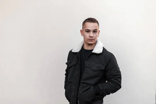 Joven guapo europeo en una chaqueta negra de moda con piel blanca en una elegante camiseta negra se encuentra cerca de la pared gris al aire libre. Modelo de chico atractivo. Calle estilo casual . — Foto de Stock