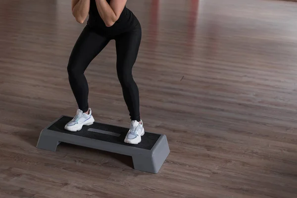 Jonge slanke vrouw in sportieve leggings in een zwarte top in de training in een fitnessruimte hurken met behulp van een platform stap. Meisjescoach toont oefeningen. Goedemorgen fitness. Close-up — Stockfoto