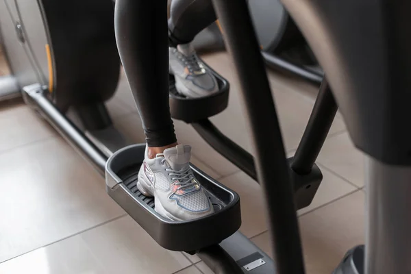 Jovem de leggings em tênis esportivos faz treinamento cardio em um simulador de passo no ginásio. A rapariga queima calorias. Fechar as pernas femininas em um simulador moderno dentro de casa . — Fotografia de Stock