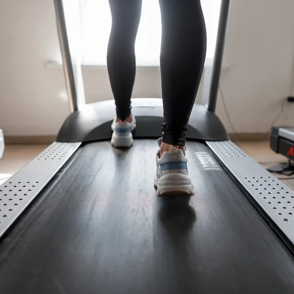 Jovem mulher em leggings negros desportivos em tênis elegantes vai em uma esteira moderna no ginásio. Menina fazendo exercício cardio para perda de peso. Bom dia ao treino. Close-up de pernas. Visão traseira . — Fotografia de Stock