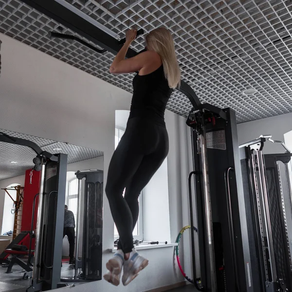 Mujer joven instructora en ropa negra con un hermoso cuerpo tira-para arriba en un simulador de metal en el gimnasio. Chica en entrenamiento en interiores. Vista trasera . —  Fotos de Stock