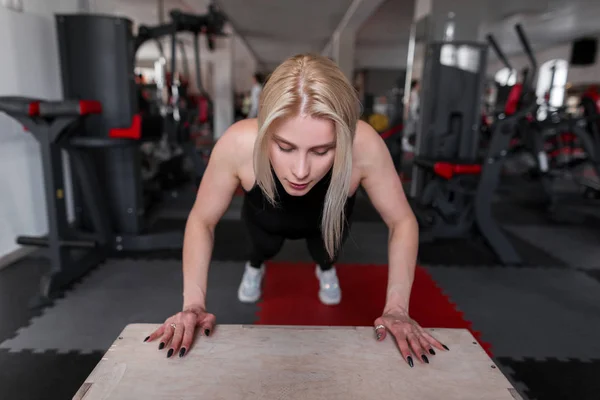 Young woman blonde in sporty black clothes is training in the gym. Girl does exercises for hands in fitness studio. Sport and beauty. — Stockfoto