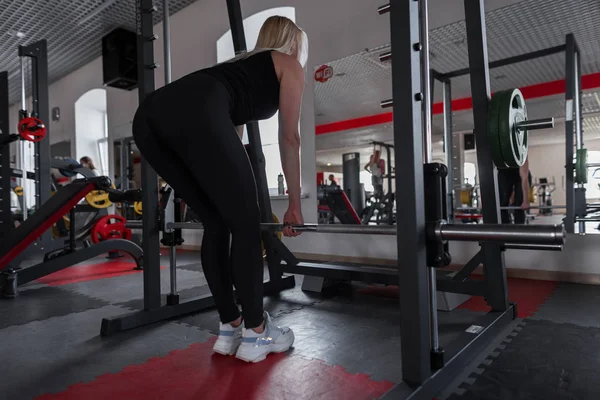Atletisk ung vacker kvinna i svart sportkläder gör övningar med en metallisk gam i ett modernt gym. Flicka tränare i utbildning. — Stockfoto