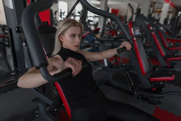 Die schöne junge Frau in schwarzer Sportbekleidung sitzt im Simulator eines Fitnessstudios. Mädchen macht die Belastung für die Hände in der Turnhalle. Vormittagstraining. — Stockfoto