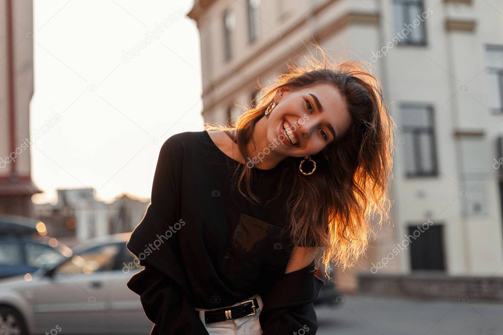 Funny positive young woman in stylish black clothes posing and smiling positively in the city on a background a spring bright orange sunset. Attractive joyful girl travels and enjoys the sunshine.