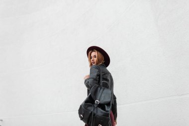 Şık bir siyah sırt çantası ile vintage deri ceket mor zarif şapka şık güzel genç kadın beyaz duvarın yakınında yürüyor. Kızıl saçlı modern kız seyahat ediyor. Bahar modası.