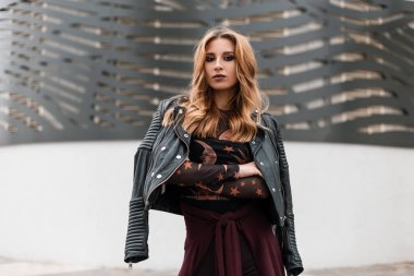 Siyah deri ceketli güzel genç kırmızı saçlı kadın, desenli şık bir bluzla şehirdeki modern binanın yakınında poz veriyor. Oldukça şık bir kız. Moda kadın giysileri. Gençlik stili.