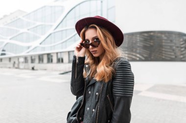 Siyah sırt çantası standları ile trendy deri ceket vintage şapka güneş gözlüğü seksi güzel genç hipster kadın standları ve kamera açık havada arıyor. Şehirli kız şehirde yürüyor. Gençlik stili.