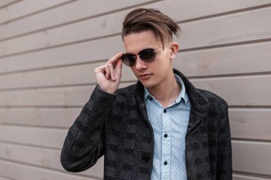Gömlek bir vintage ekose ceket şık güneş gözlüğü trendy saç modeli ile Çekici genç hipster adam ahşap bir duvarın yakınında sokakta pozlar. Sevimli adam şehirde açık havada. Modern erkek giyim